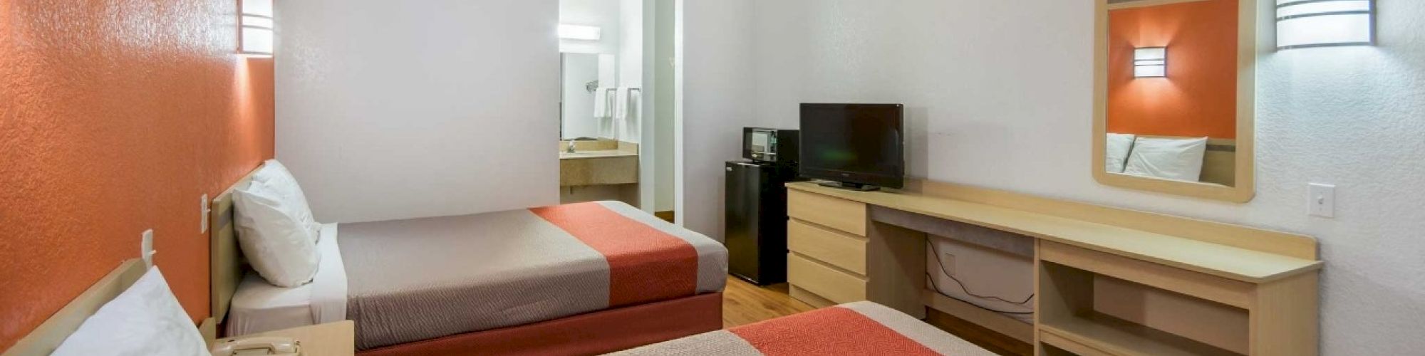 This image shows a hotel room with two beds, an orange accent wall, a dresser with a TV, a mirror, and a small bathroom in the background.