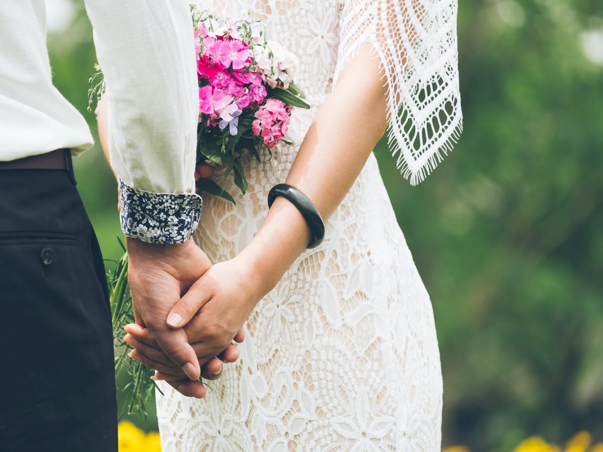 A couple is holding hands, with the bride wearing a white lace dress and holding a bouquet of pink flowers. They appear to be at a wedding.