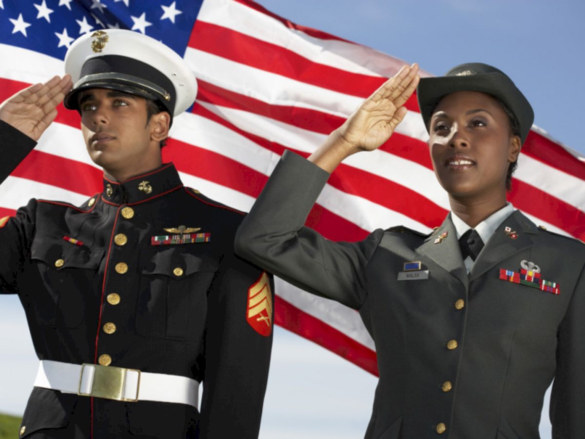 Two military personnel in uniform salute in front of an American flag.