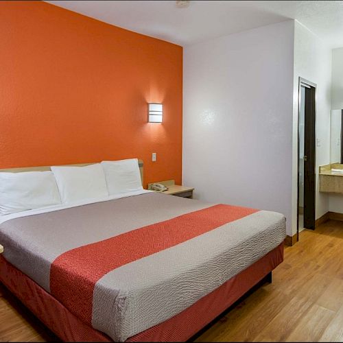 This image shows a hotel room with a king-size bed, orange and white walls, bedside tables, a desk, chair, and a sink area with a mirror.