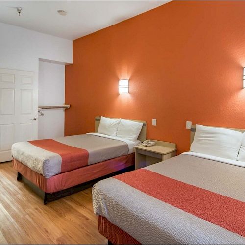 A hotel room with two beds, orange accent wall, wooden flooring, bedside tables, wall sconces, and a view into the bathroom.