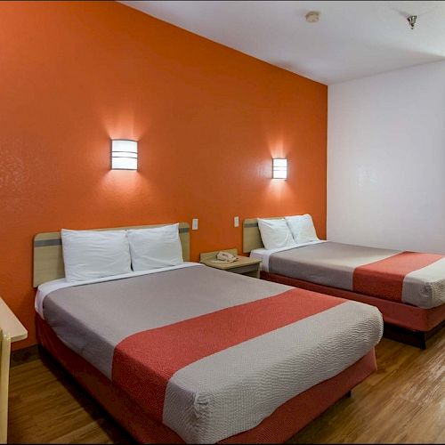 The image shows a hotel room with two double beds, orange accent wall, a table with chairs, and a vanity area with a sink and mirror.