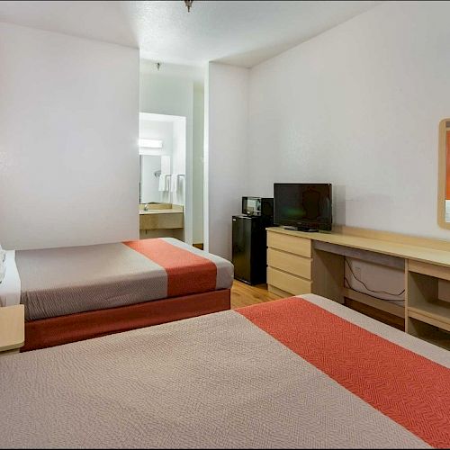 The image shows a hotel room with two beds, an orange accent wall, a TV on a dresser, and a desk with a mirror. There is a bathroom in the background.