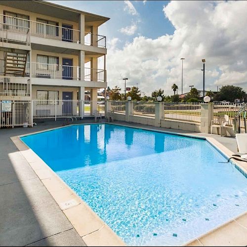 This image shows an outdoor pool area beside a multi-story building with balconies, a patio chair, and a table, all under a partially cloudy sky.