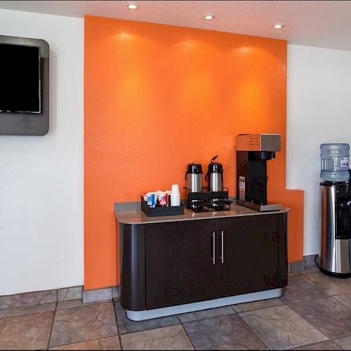 The image shows a small kitchenette within a room, featuring an orange accent wall, a flat-screen TV, a coffee station, and a water dispenser.