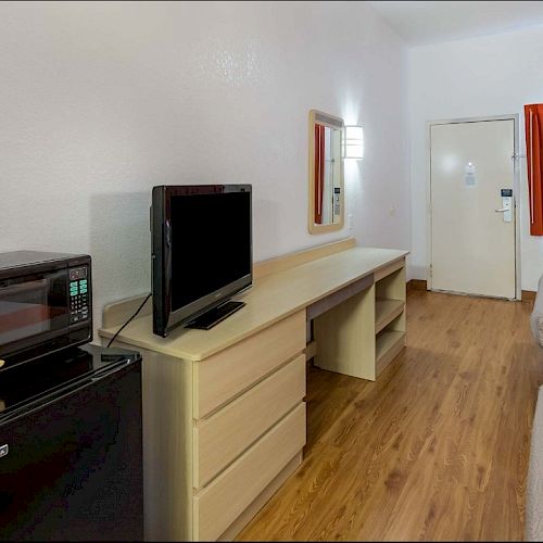 The image shows a hotel room with two beds, a TV on a dresser, a microwave, a mini-fridge, a desk, and a window with red curtains ending the sentence.