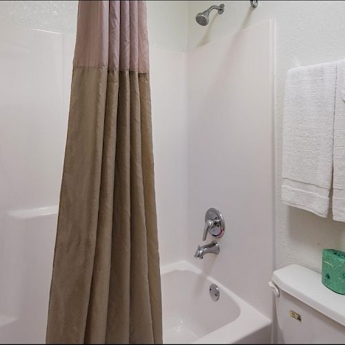 The image shows a bathroom with a shower-tub combo, a beige shower curtain, white towels on a rack, a green container, and a toilet with a silver handle.