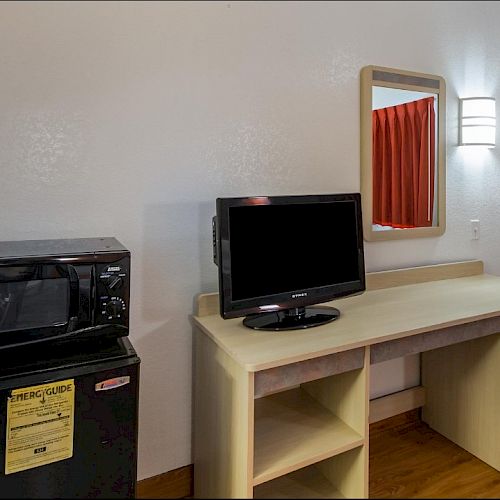 The image shows a hotel room with a TV on a desk, a microwave on a mini-fridge, a mirror, and a light fixture above the desk.