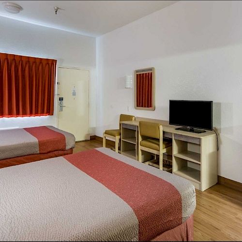 The image shows a hotel room with two beds, a TV on a desk, chairs, a mini-fridge, and a window with red curtains. The decor is simple and modern.