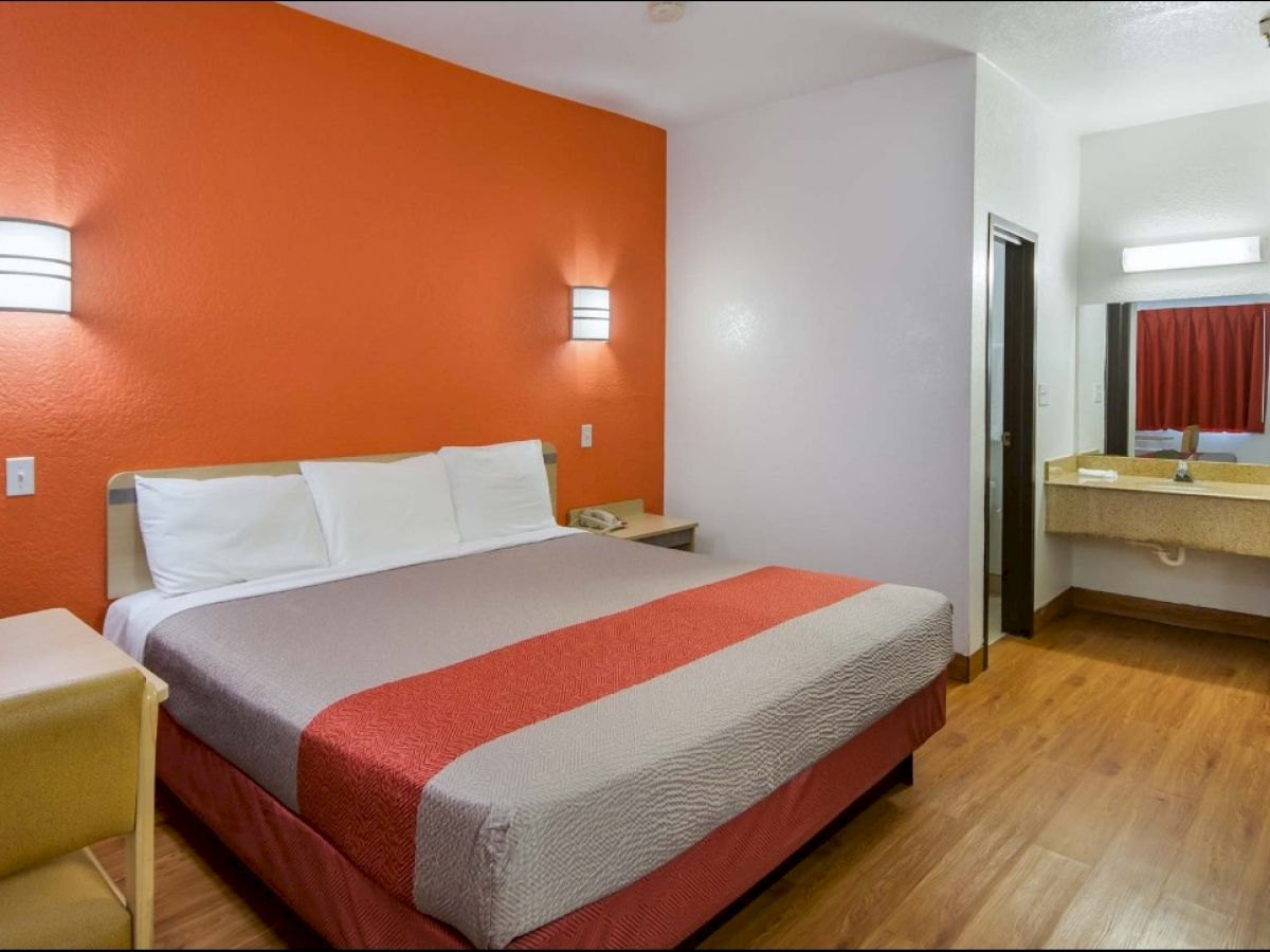 A modern hotel room with a king-sized bed, orange accent wall, bedside lights, a small table, and a vanity area with a sink and mirror.