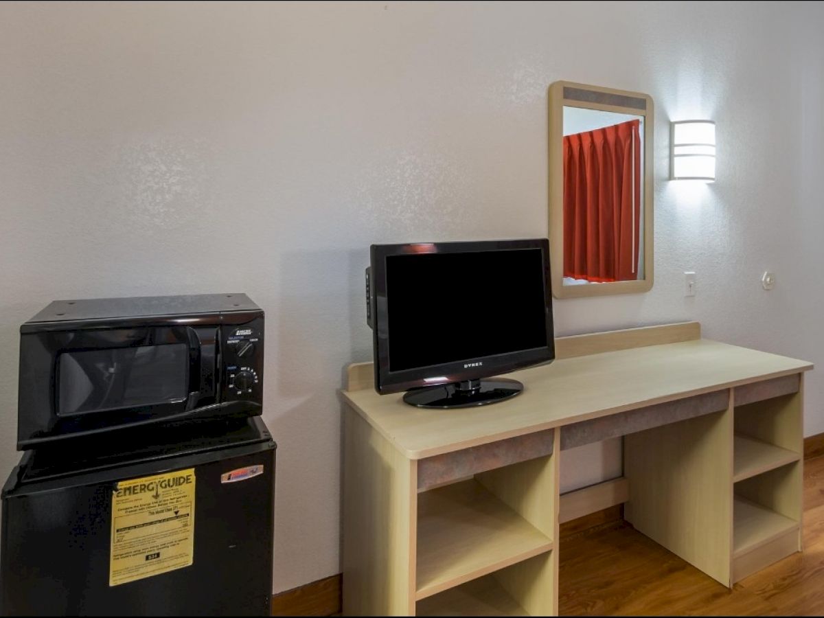 The image shows a small room setup with a microwave and mini-fridge on the left, a TV on a desk with shelves underneath, a mirror and a light on the wall.