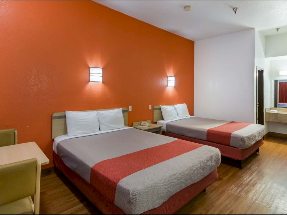 A hotel room with two double beds, orange accent wall, wall lights, a table with chairs, a dresser, and a sink area with a mirror in the background.