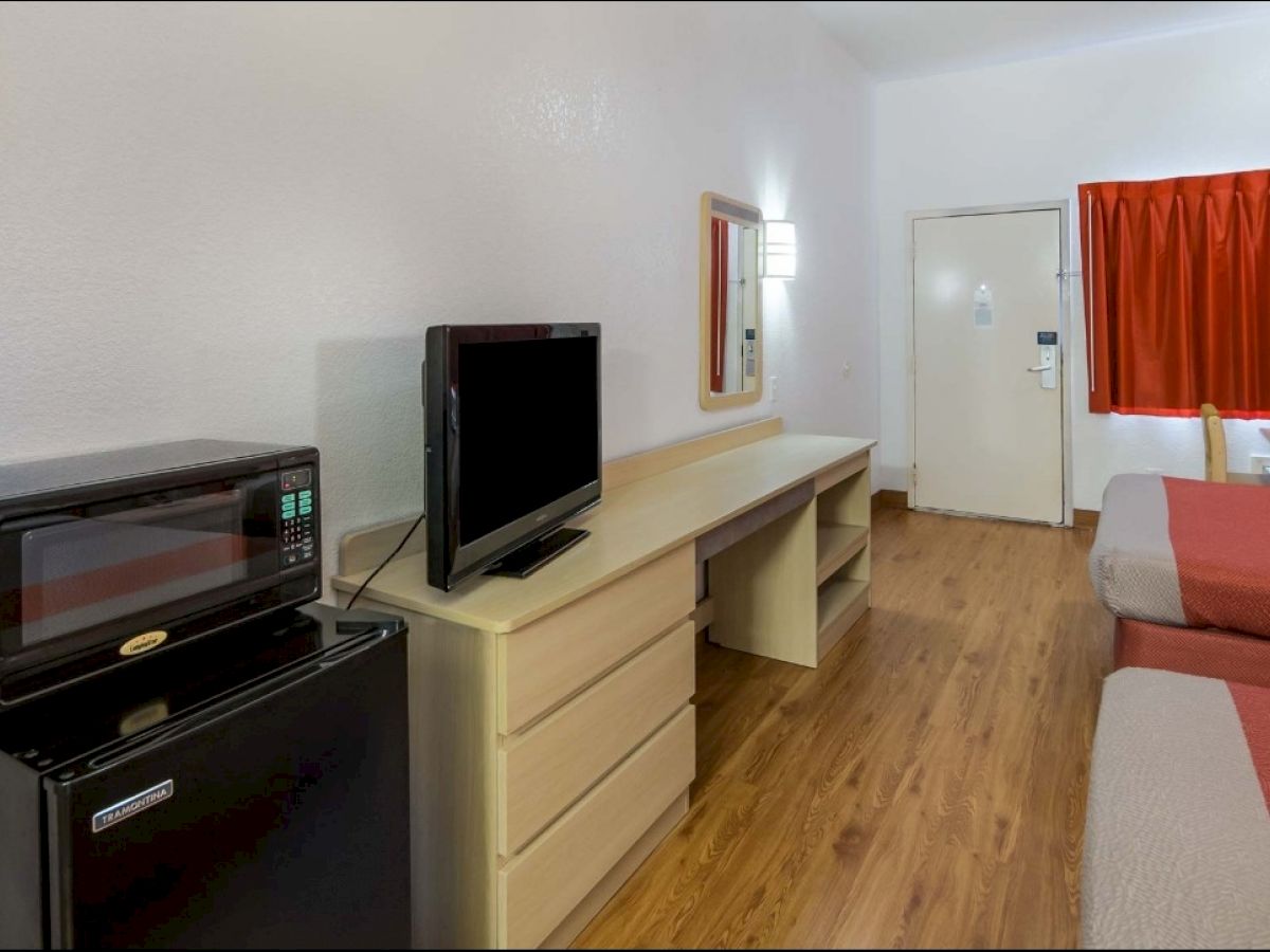 The image shows a hotel room with two beds, a desk, a TV, a microwave, a mini fridge, and a window with red curtains, all on a wooden floor.