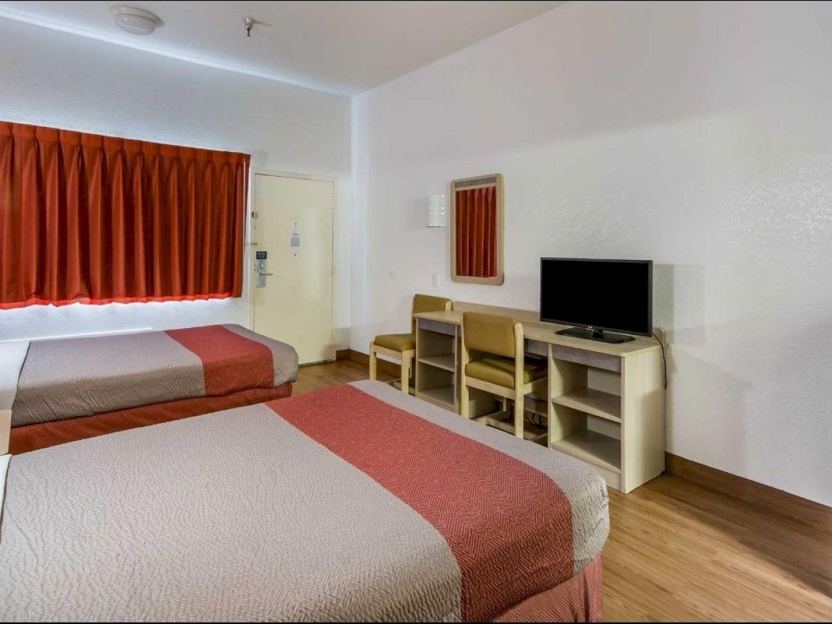 The image shows a hotel room with two beds, a TV on a desk with chairs, a mini-fridge, and a window with red curtains.