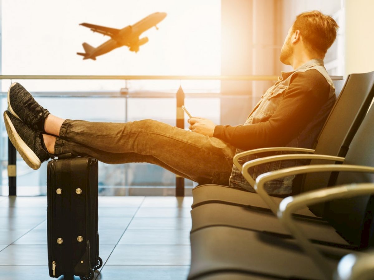 A person is sitting in an airport lounge with their feet on a suitcase, looking at a plane taking off through the window.