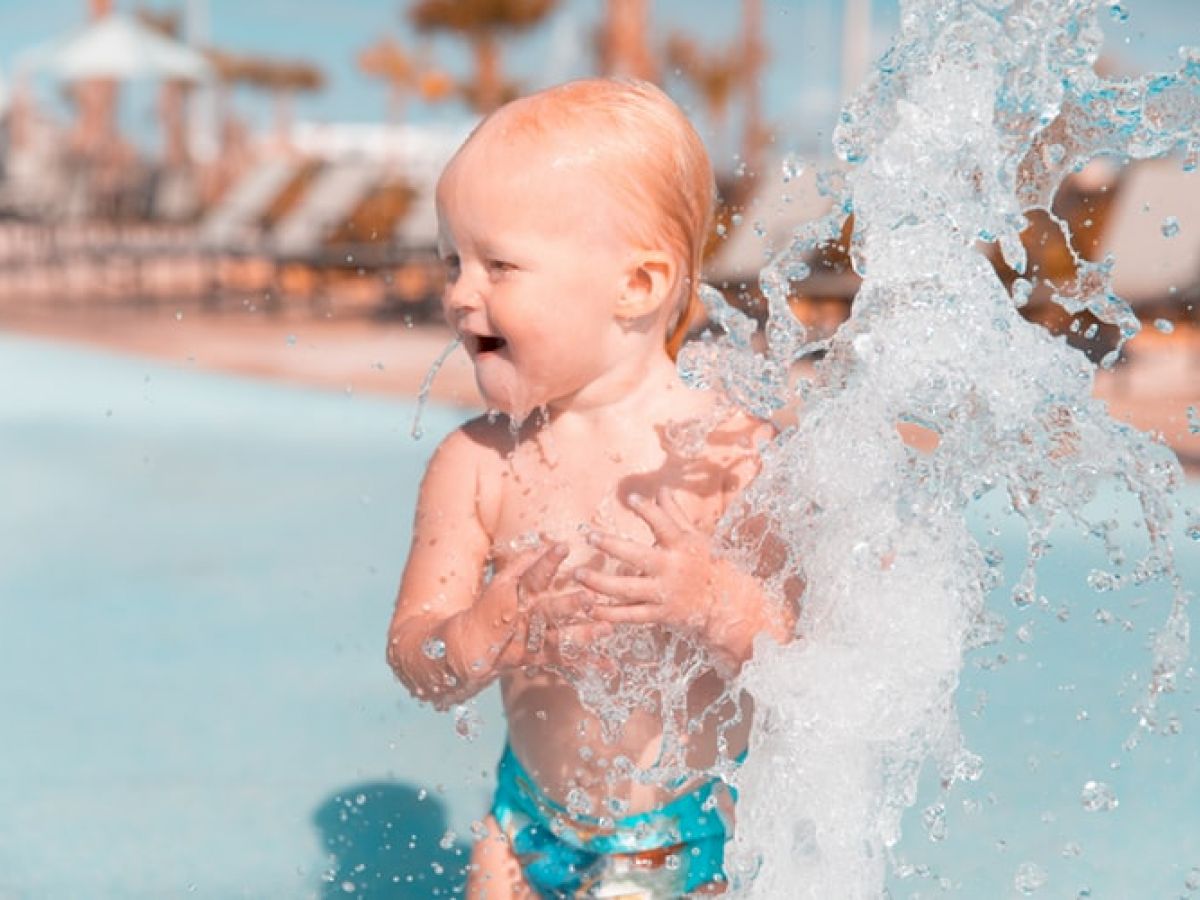 A toddler wearing a blue swimsuit is enjoying playing with water at a pool, with water splashing and a sunlit background of poolside furniture.