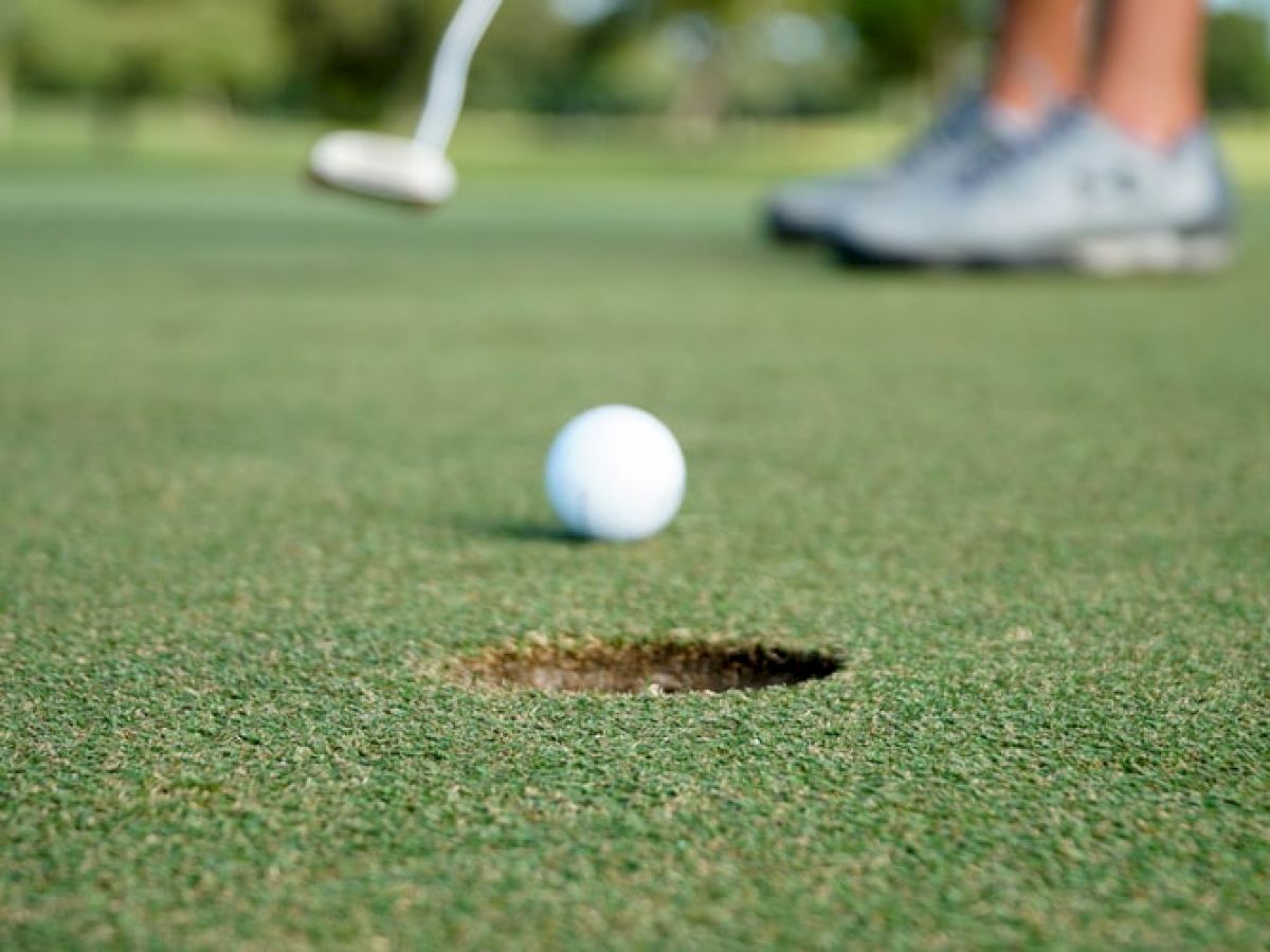 A golf ball is close to a hole on a putting green, with a golfer's legs and putter visible in the background.