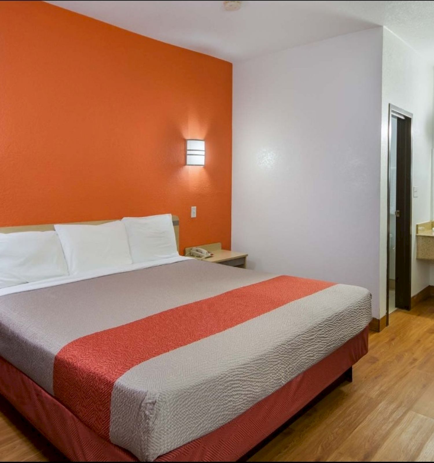 The image shows a modern, minimal hotel room with a double bed, orange accent wall, wall lights, wood flooring, a table, a sink, and a mirror area.