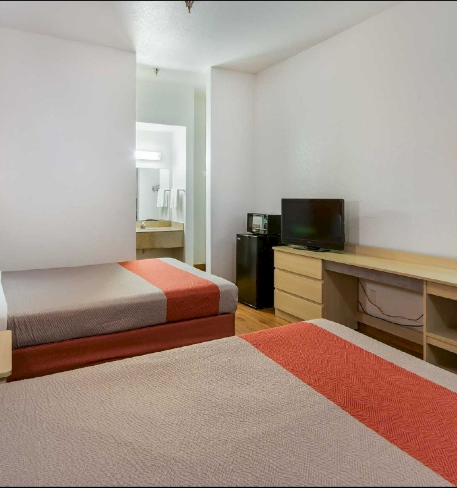 A hotel room with two beds, a TV, a dresser, a desk, and a small washroom. The room has orange and beige decor with simple furnishings.