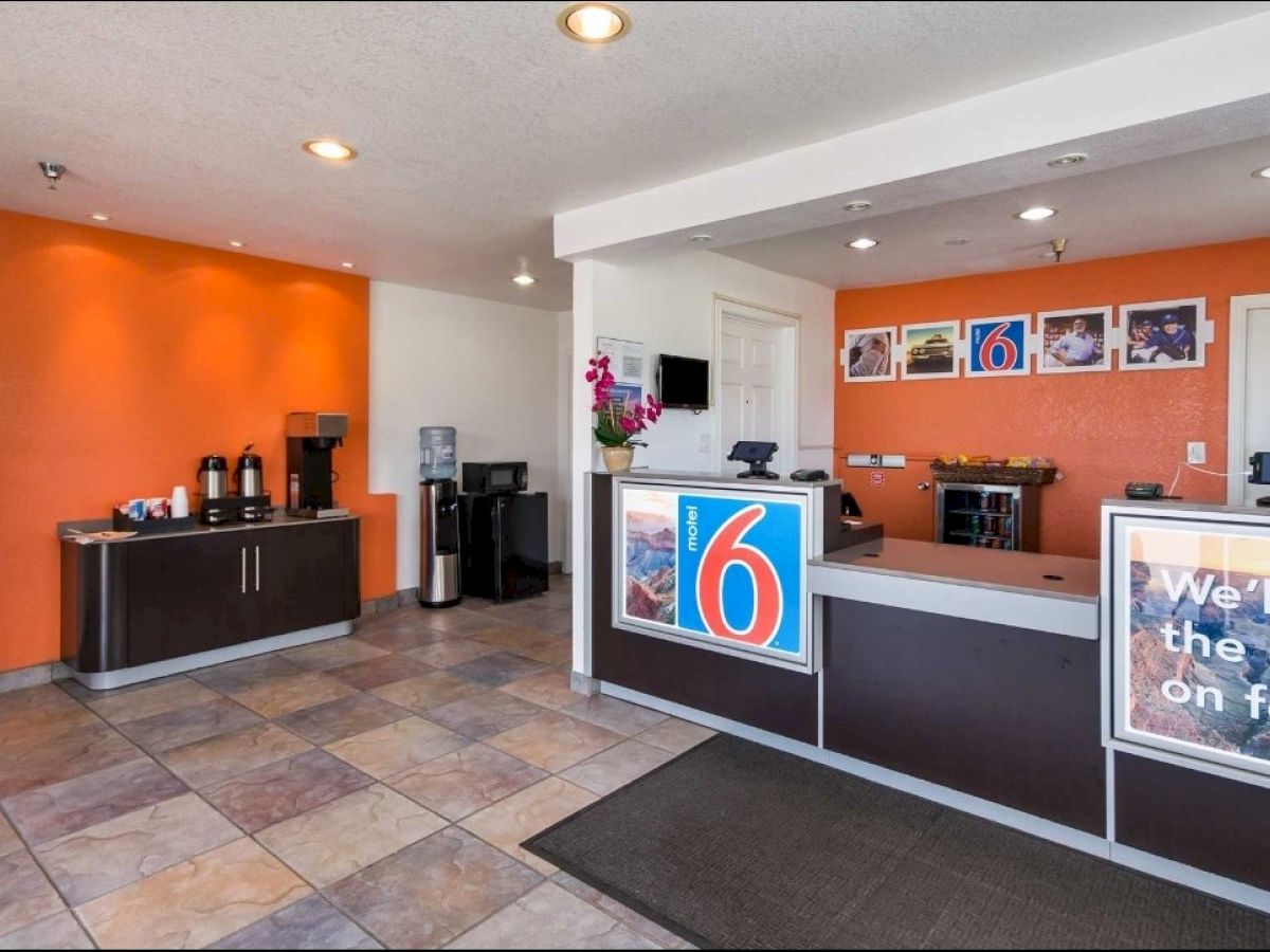 This image shows a hotel lobby with a reception desk, coffee station, and orange accent wall, featuring Motel 6 branding and the slogan 