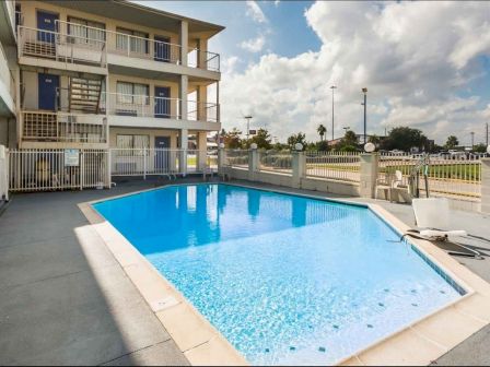 The image shows an outdoor swimming pool with a building and parking lot in the background. Lounge chairs and a fence surround the pool area.