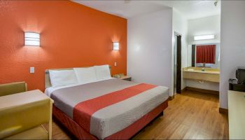 A hotel room with an orange accent wall, a bed, a small sofa, a table, and a connected bathroom with a sink and a mirror is shown.