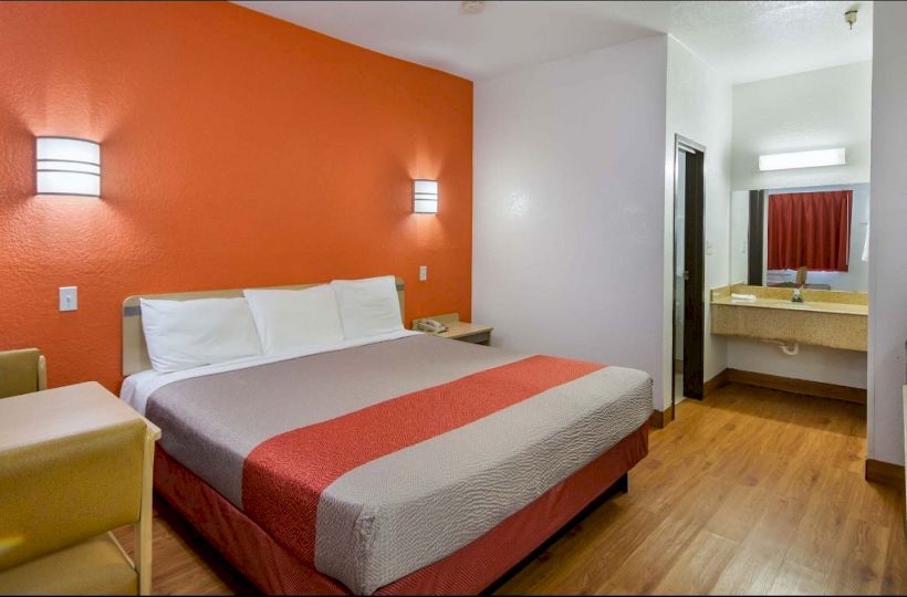 A hotel room with an orange accent wall, a bed, a small sofa, a table, and a connected bathroom with a sink and a mirror is shown.