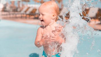 A young child in swimwear happily plays with water from a splash fountain at a poolside, enjoying the sunny day.