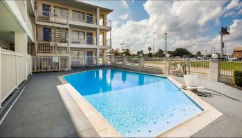 The image shows an outdoor pool area with lounge chairs, next to a multi-story building, under a partly cloudy sky.