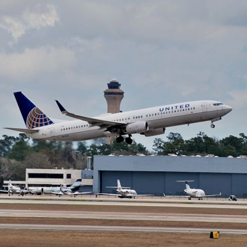 An airplane is taking off from an airport runway, with a control tower and other aircraft visible in the background.