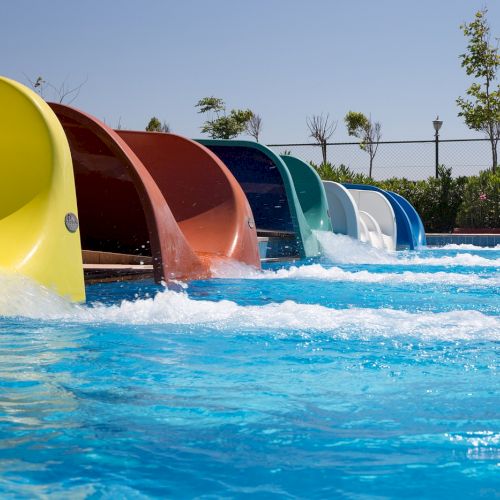 The image shows a pool with multiple colorful water slides side by side, with trees and a fence in the background under a clear sky.