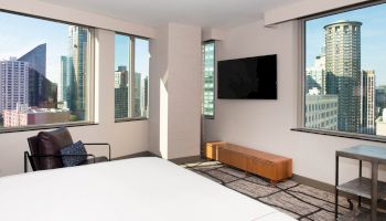 This image showcases a modern hotel room with city views, featuring large windows, a wall-mounted TV, and contemporary furnishings.