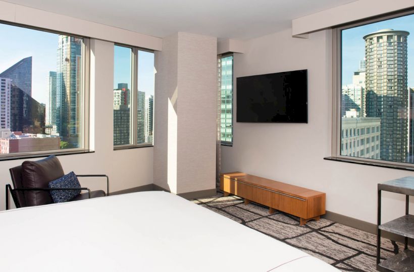 This image showcases a modern hotel room with city views, featuring large windows, a wall-mounted TV, and contemporary furnishings.