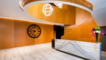 A modern lobby with a marble reception desk, wooden walls, a large clock, spiral staircase, and decorative lighting. Purple flowers on the desk.