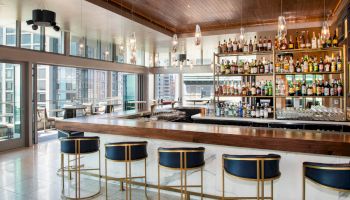 The image shows a modern bar with high stools, a well-stocked liquor shelf, and large windows offering plenty of natural light and city views.