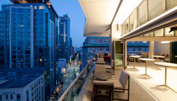 The image shows a modern balcony with seating areas overlooking a cityscape at dusk, featuring tall buildings and a bustling urban street below.
