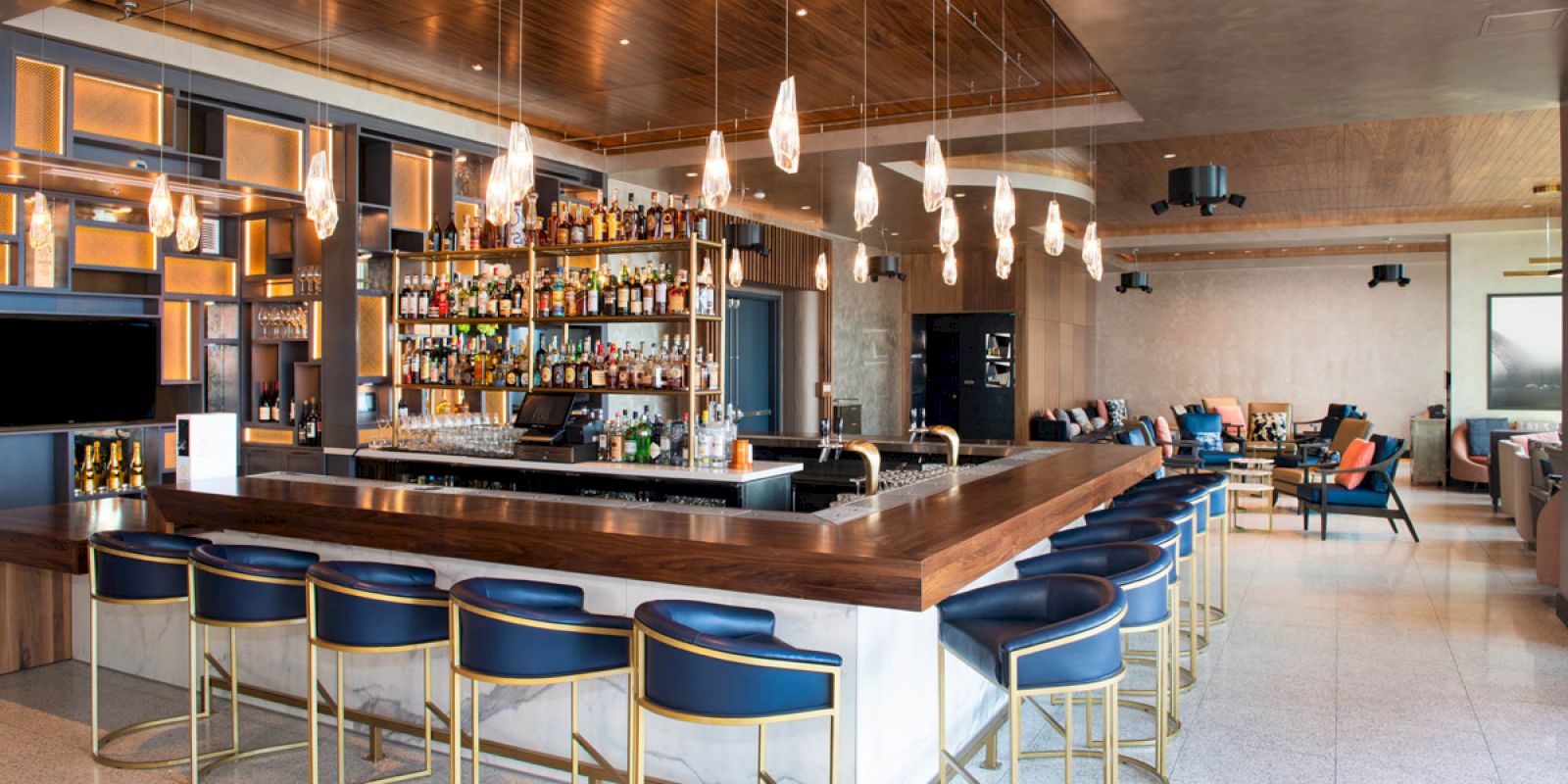 The image shows a modern bar with a wooden countertop, bar stools, hanging lights, and a well-stocked liquor shelf in a spacious lounge area.
