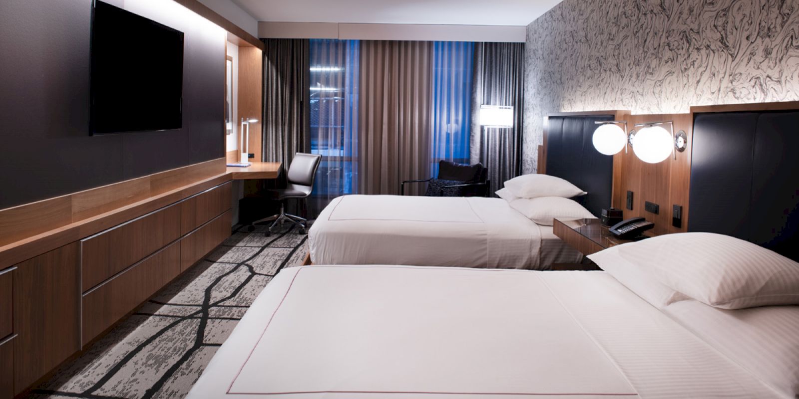 A hotel room with two neatly made beds, a wall-mounted TV, a desk area, a chair, and large windows with curtains.