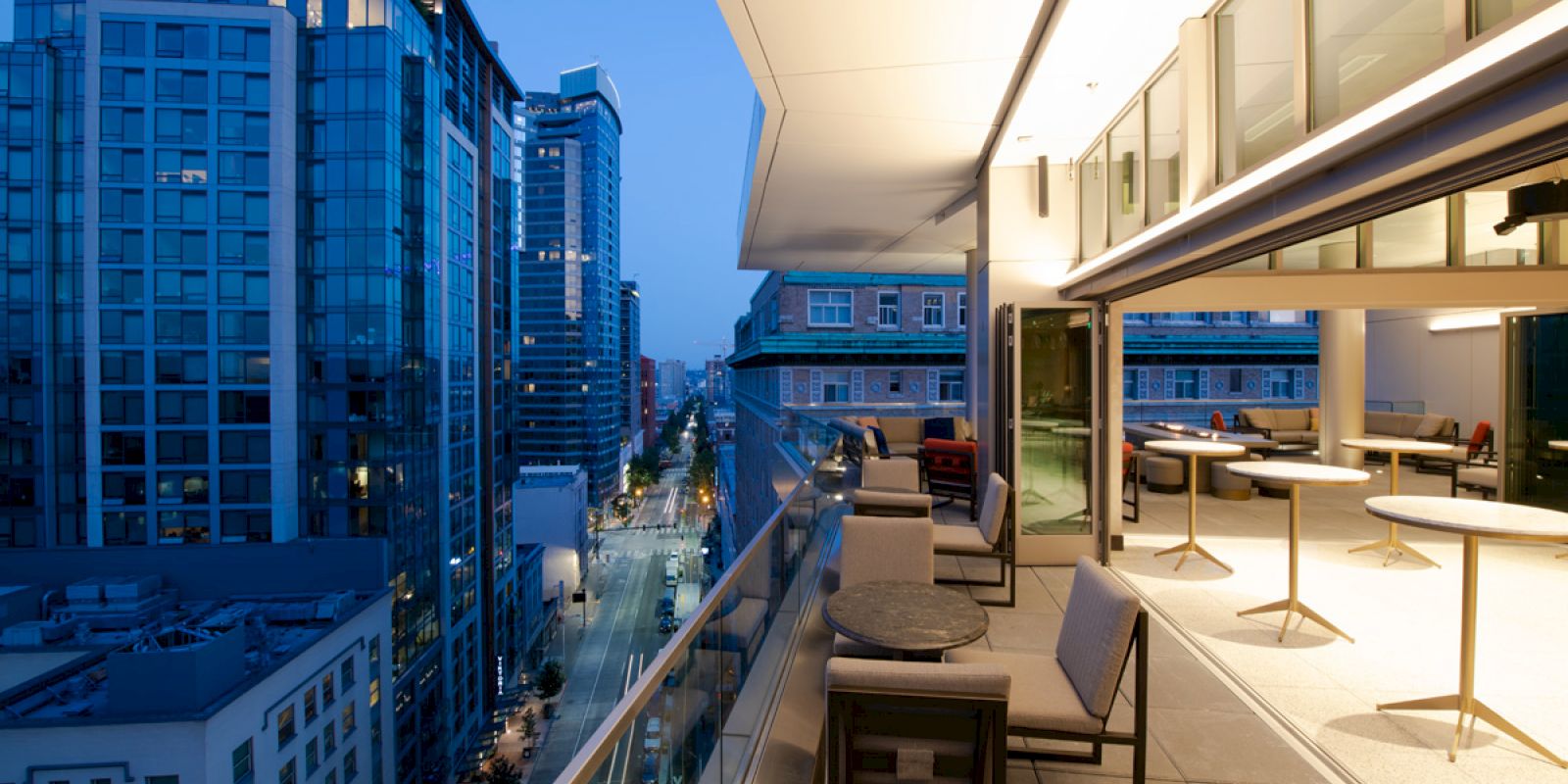 A modern cityscape at dusk with tall buildings and a stylish balcony featuring seating and tables overlooking the urban street below.