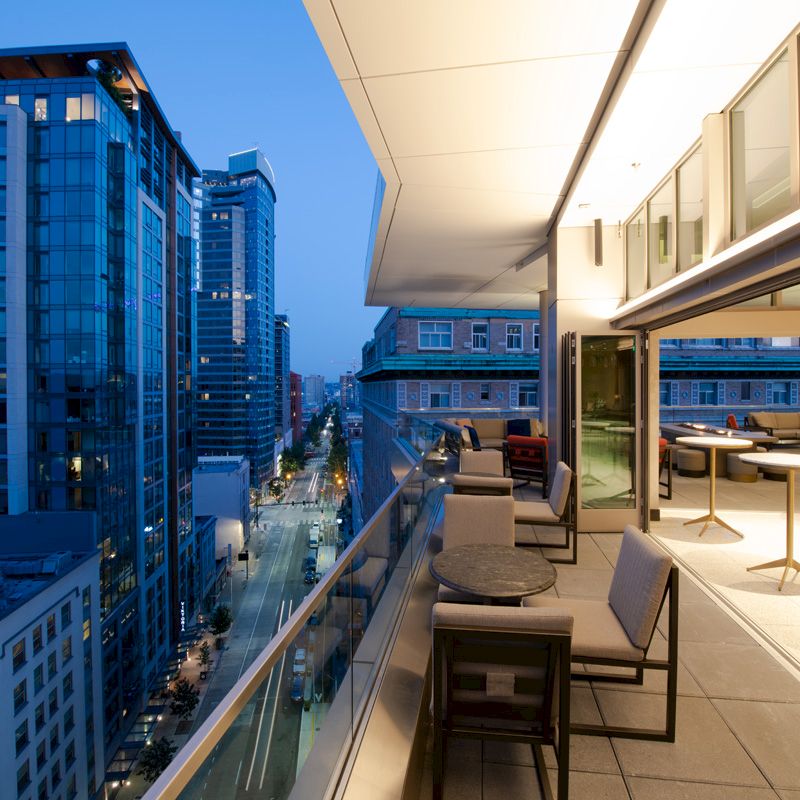 A cityscape at dusk featuring a modern high-rise terrace with tables and chairs on the right, overlooking a street lined with tall buildings.