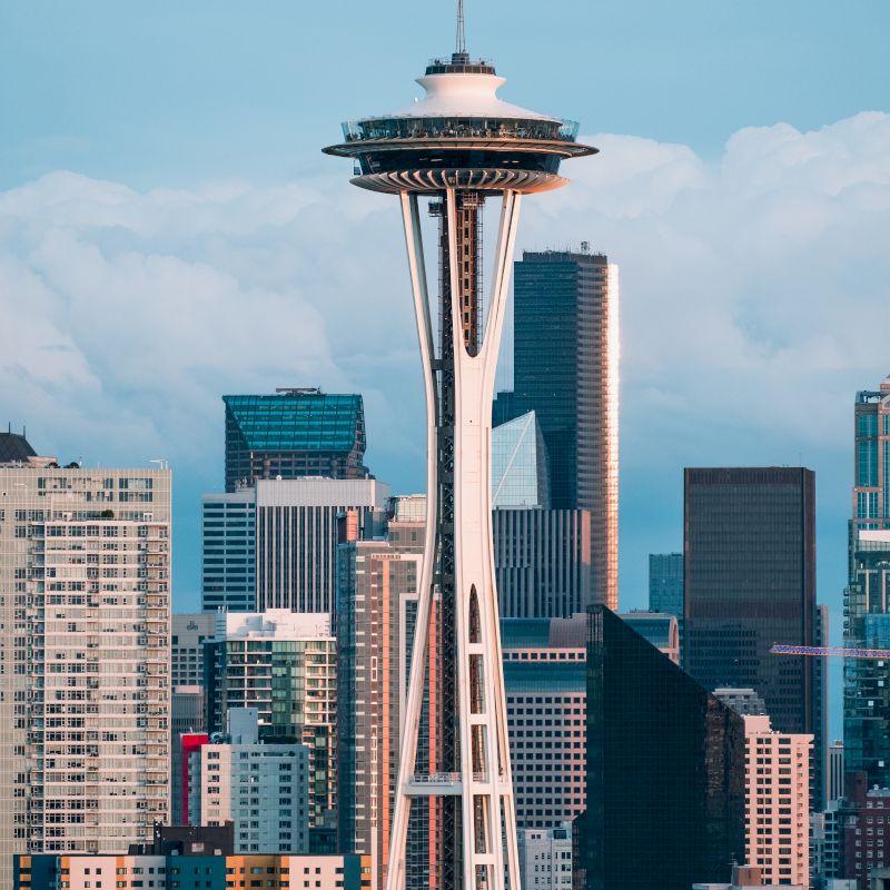 The image shows the Seattle Space Needle with a backdrop of various downtown skyscrapers and buildings under a partly cloudy sky.