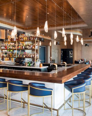 A stylish modern bar with blue barstools, a wooden countertop, shelves filled with various bottles, pendant lights, and a cozy seating area.