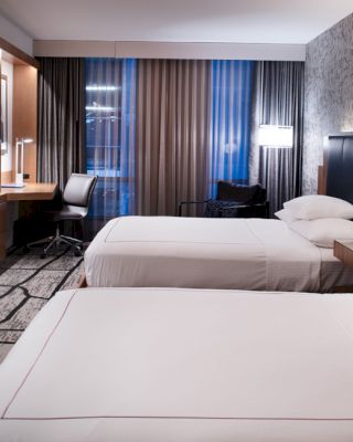 A modern hotel room with two beds, a wall-mounted TV, a desk with a chair, and patterned curtains, appears neat and inviting, ending the sentence.