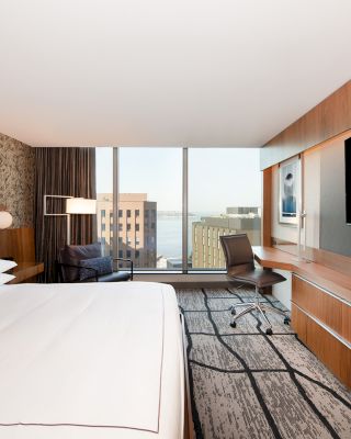 A modern hotel room with a large bed, desk, chair, wall-mounted TV, and a window showing a cityscape and water view in the background.