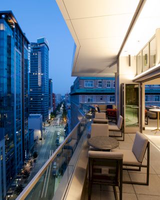 A rooftop terrace with seating and tables overlooks a cityscape with tall buildings and a street at dusk.