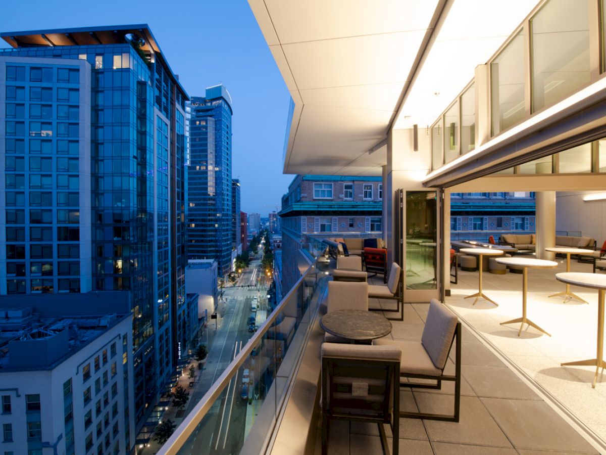 A modern balcony at dusk overlooks a city street, with outdoor seating and tables along the side. The skyline features tall buildings.