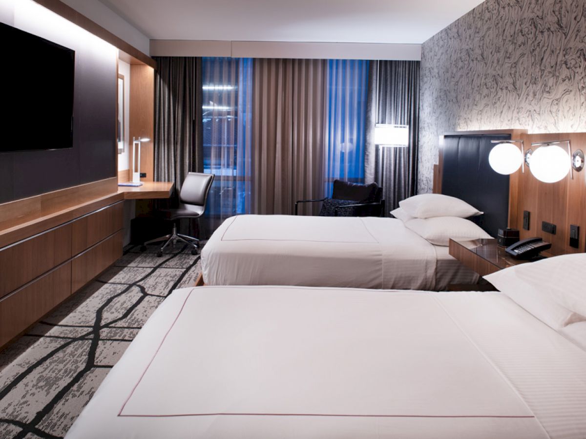 The image shows a modern hotel room with two beds, a desk, a chair, a TV, and a window with curtains. The decor is contemporary with subtle lighting.