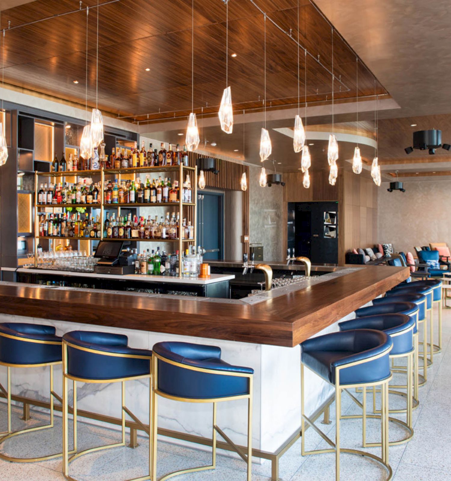The image shows a modern bar with a wooden counter, blue chairs, various bottles on shelves, and hanging pendant lights, within a spacious lounge area.