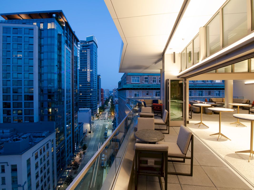 A modern high-rise balcony view at dusk, overseeing a cityscape with tall buildings and a neatly arranged outdoor seating area.