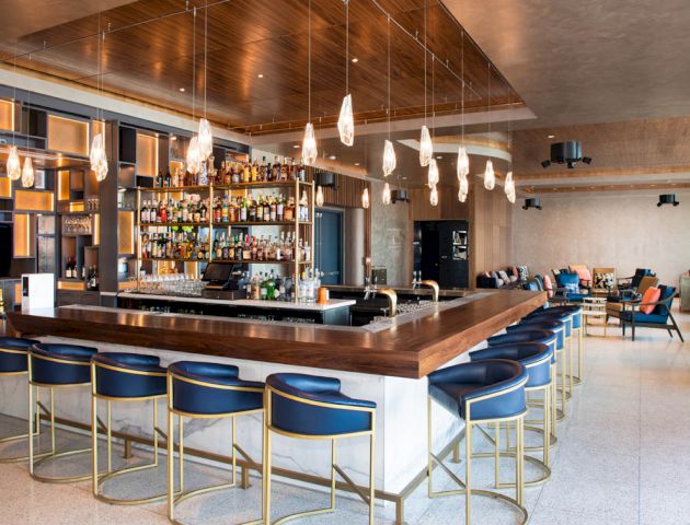 This image features a modern bar area with a wooden countertop, a variety of hanging lights, cushioned bar stools, and a well-stocked liquor display.